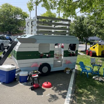 Children's playhouse designed to look like a travel trailer, with plastic chairs and a grill set up to look like a camping trip