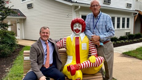 Two men sitting and standing by a Ronald McDonald statue.