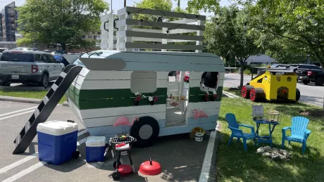 Children's playhouse designed to look like a travel trailer, with plastic chairs and a grill set up to look like a camping trip