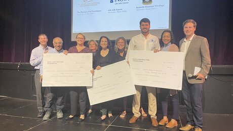 A group of people holding three large $20,000 checks, made out to non-profits: The Michael J. Fox Foundation for Parkinson’s Research, INOVA Life with Cancer, and Rockville Volunteer Fire Department.