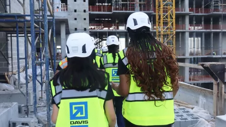 Two women walk on a construction site in the foreground. Others are in the background