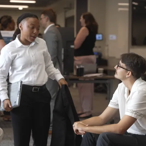 Two interns speak to one another. One sits on a couch, the other stands.
