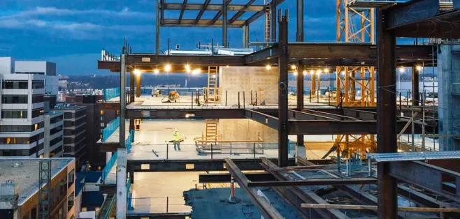 Multi-story building under construction at dusk, with exposed steel framing, poured concrete floors lit by work lights, a tower crane, and a hard-hatted worker on an open deck above the city skyline.