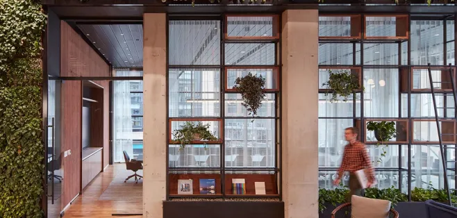 Modern office interior with a floor-to-ceiling living green wall framing a glass-walled conference room, warm wood floors, and an employee walking past holding a laptop.
